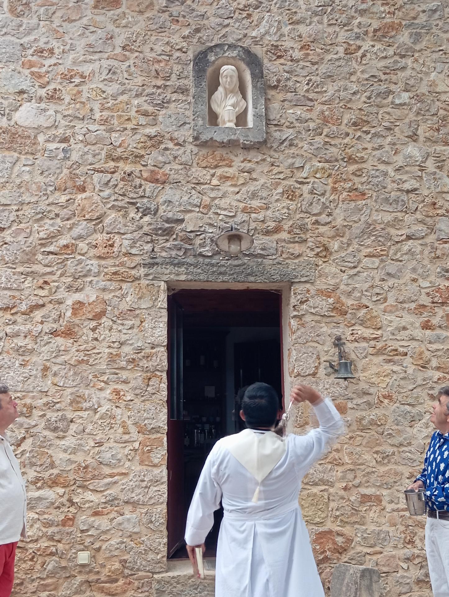 La bénédiction du buste de sainte Marie-Madeleine : le prêtre en aube bénit l'œuvre au goupillon dans sa niche, au-dessus de la porte, au Domaine Les Terres Promises, le 19 juillet 2025 — trois jours avant la fête de la sainte. Sculpteur d'art sacré Jean-Joseph Chevalier (Brignoles, Var, Provence).