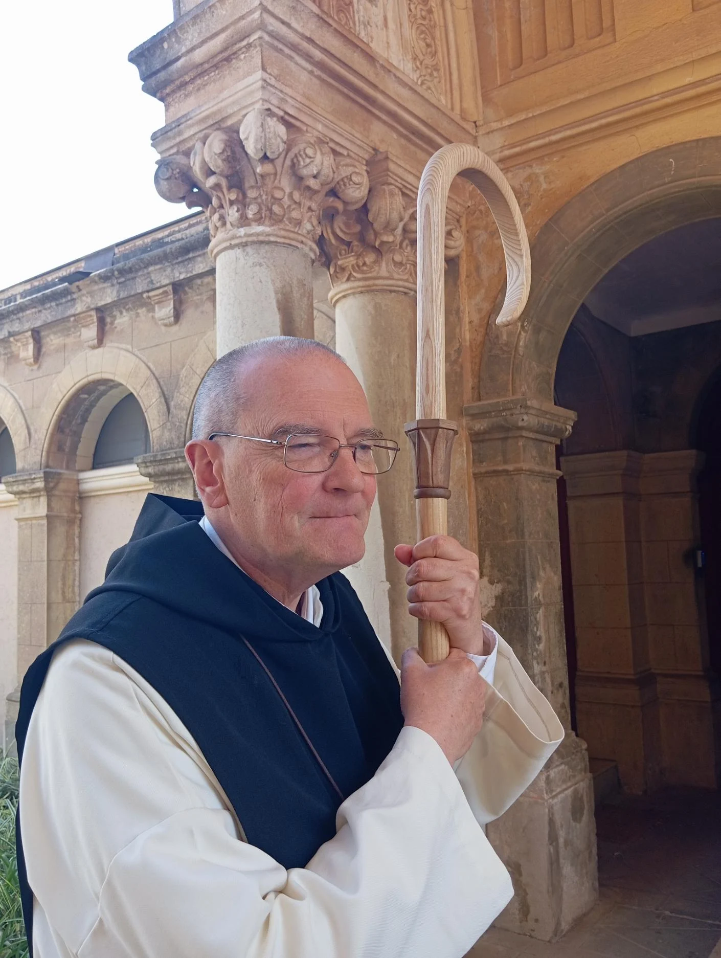 Dom Jean-Marie Gervais, abbé de Notre-Dame de Lérins, tenant sa crosse abbatiale en frêne et noyer sculptée par Jean-Joseph Chevalier — portrait dans le cloître de l'abbaye après la bénédiction abbatiale du 2 mai 2026, Congrégation Cistercienne de l'Immaculée Conception, ancien prieur de Sénanque 1997-2025