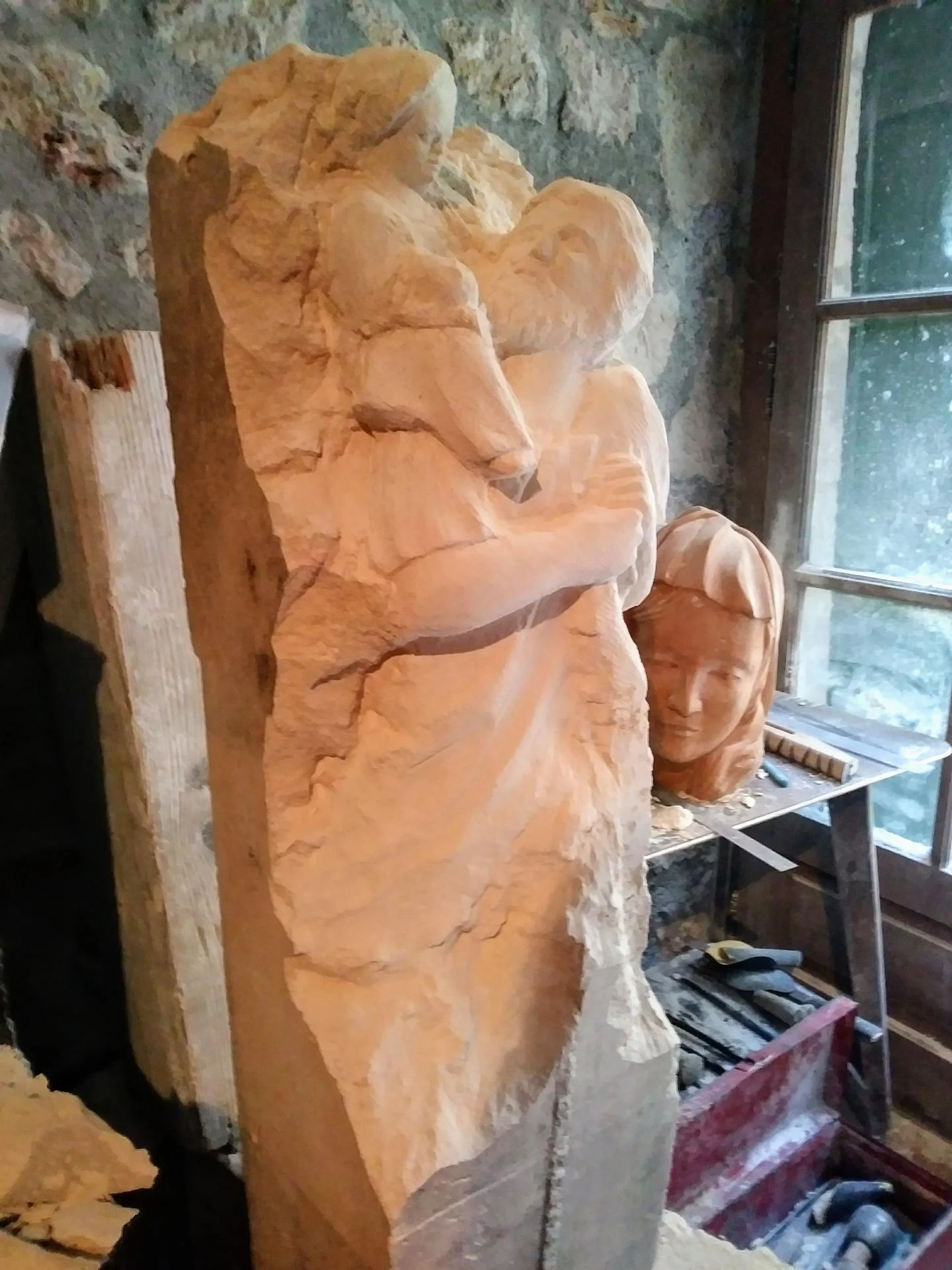Vue d'atelier — la statue de saint Christophe en cours de taille directe, émergeant progressivement du bloc de pierre calcaire naturelle devant la fenêtre de l'atelier. Le visage du saint modelé en terre cuite est posé à côté comme référence. Sans moulage ni report mécanique : l'œuvre est arrachée au bloc geste après geste, technique traditionnelle de la taille directe à la main. Sculpteur d'art sacré chrétien Jean-Joseph Chevalier, atelier de Brignoles (Var, Provence). Commande d'un particulier, 2017.