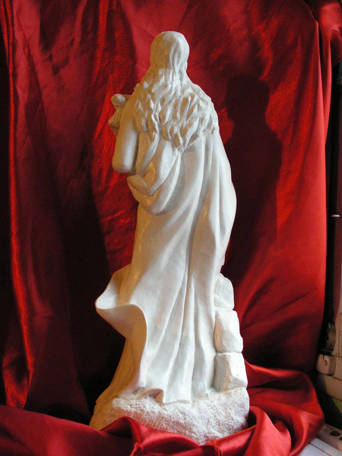 Statue Sainte Philomène vue de dos — chevelure dénouée tombant en longues mèches sculptées une à une dans le vent, cape en mouvement, ronde-bosse complète. Pierre calcaire naturelle blanche 70 cm, taille directe complète, finition à la gradine jusque dans les replis du dos — opposé à la statuaire en demi-bosse industrielle adossée au mur. Atelier du sculpteur sur pierre d'art sacré chrétien Jean-Joseph Chevalier (Brignoles, Var). Cadeau d'ordination sacerdotale 2008.
