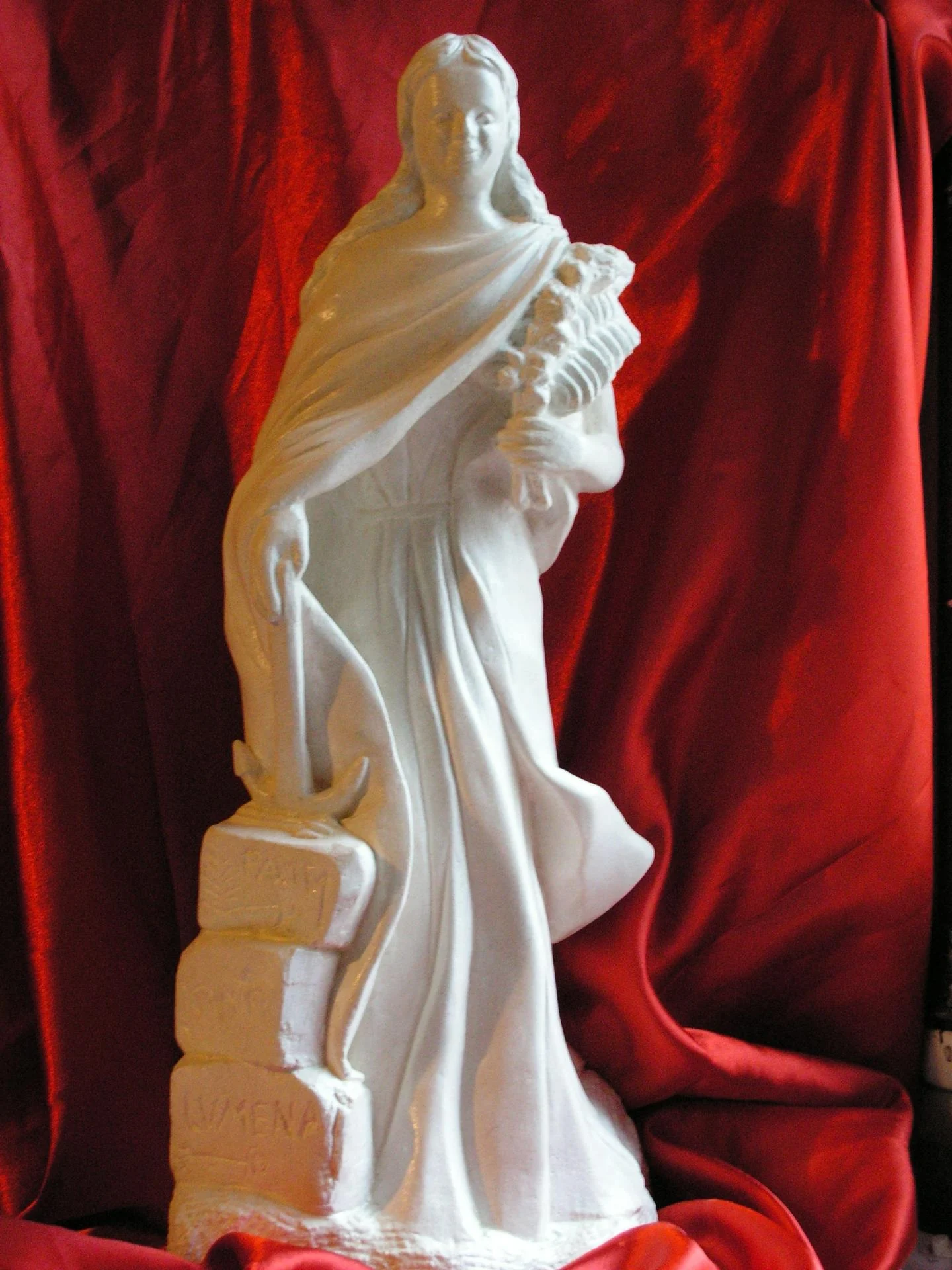 Statue Sainte Philomène vue de face — sculpture religieuse en pierre naturelle calcaire blanche 70 cm, taille directe à la main par le sculpteur d'art sacré chrétien Jean-Joseph Chevalier. La jeune vierge et martyre debout, drapé emporté par le vent, ancre marine du martyre à ses pieds, palme du martyre et gerbe de fleurs de lys de la virginité dans la main gauche, épitaphe LUMENA PAX TE CUM FI gravée en lettres capitales antiques sur les trois pierres du socle, transcription exacte de la dalle de la catacombe de Priscille à Rome (25 mai 1802). Cadeau d'ordination sacerdotale 2008, statue de dévotion privée pour oratoire de prêtre.