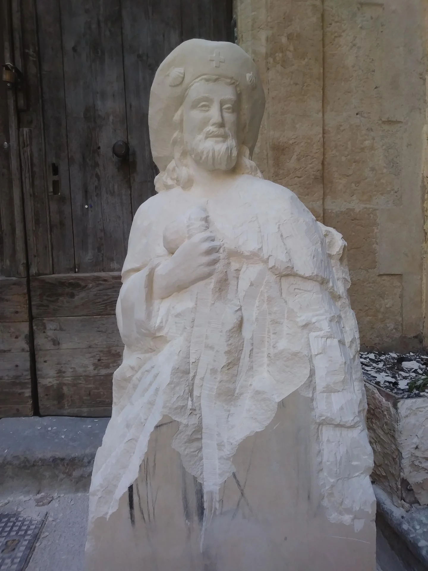Statue Saint Jacques de Compostelle en cours de dégrossissage à la pointe et à la gradine — la silhouette du pèlerin, son chapeau à large bord et les premiers plis du manteau émergent du bloc de pierre calcaire encore en partie brut. Taille directe à la main pour l'abbaye Saint-Paul de Wisques. Sculpture saint Jacques pierre naturelle France, étape de modelage en pierre