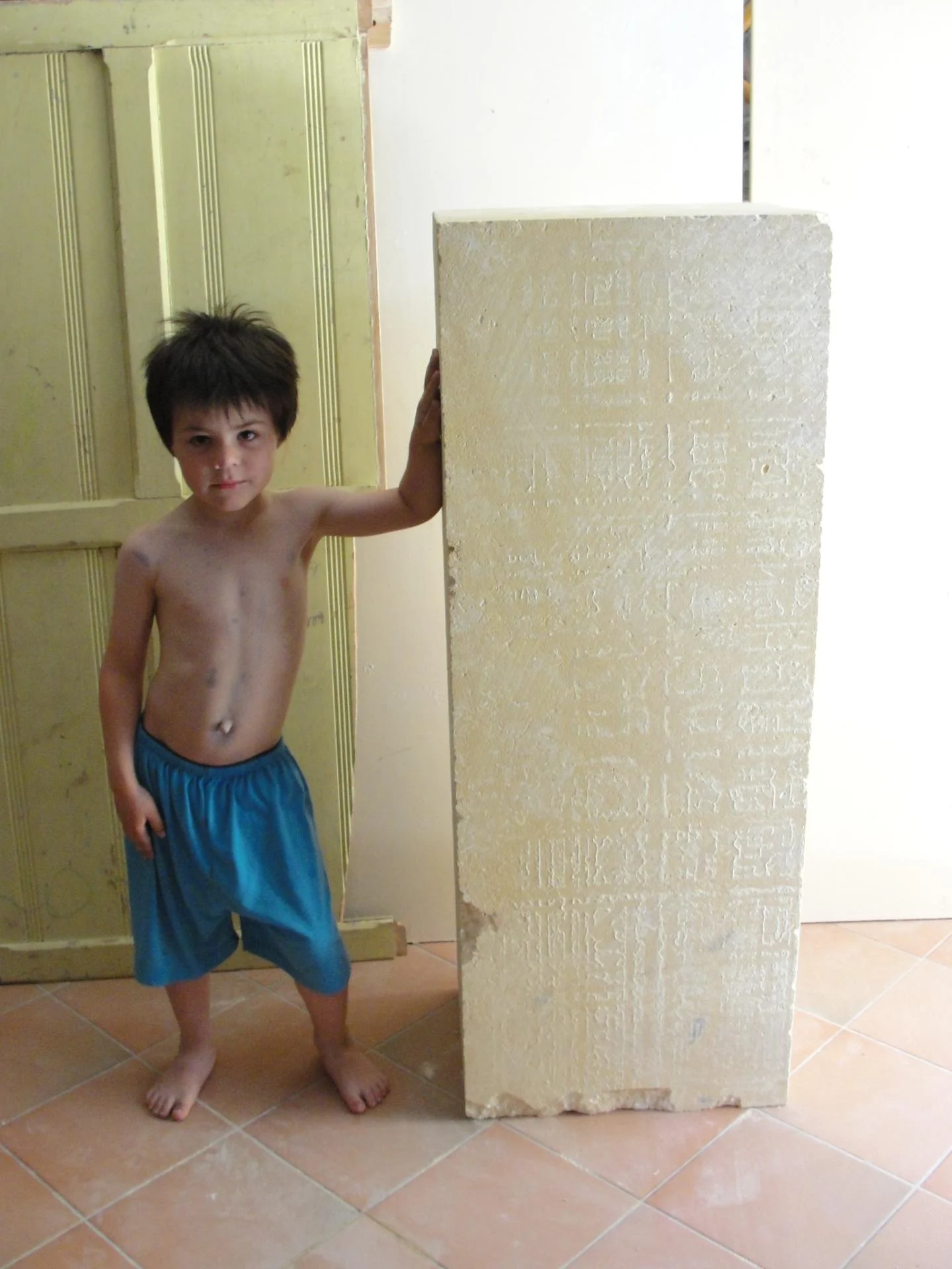 L'atelier — le bloc capable de la statue Saint Jean dressé près d'un enfant qui en donne l'échelle humaine. Pierre naturelle 1,20 m, taille directe. Ensemble des quatre évangélistes pour l'abbaye de Lagrasse 2016 — Jean-Joseph Chevalier, sculpteur d'art sacré chrétien (Var).