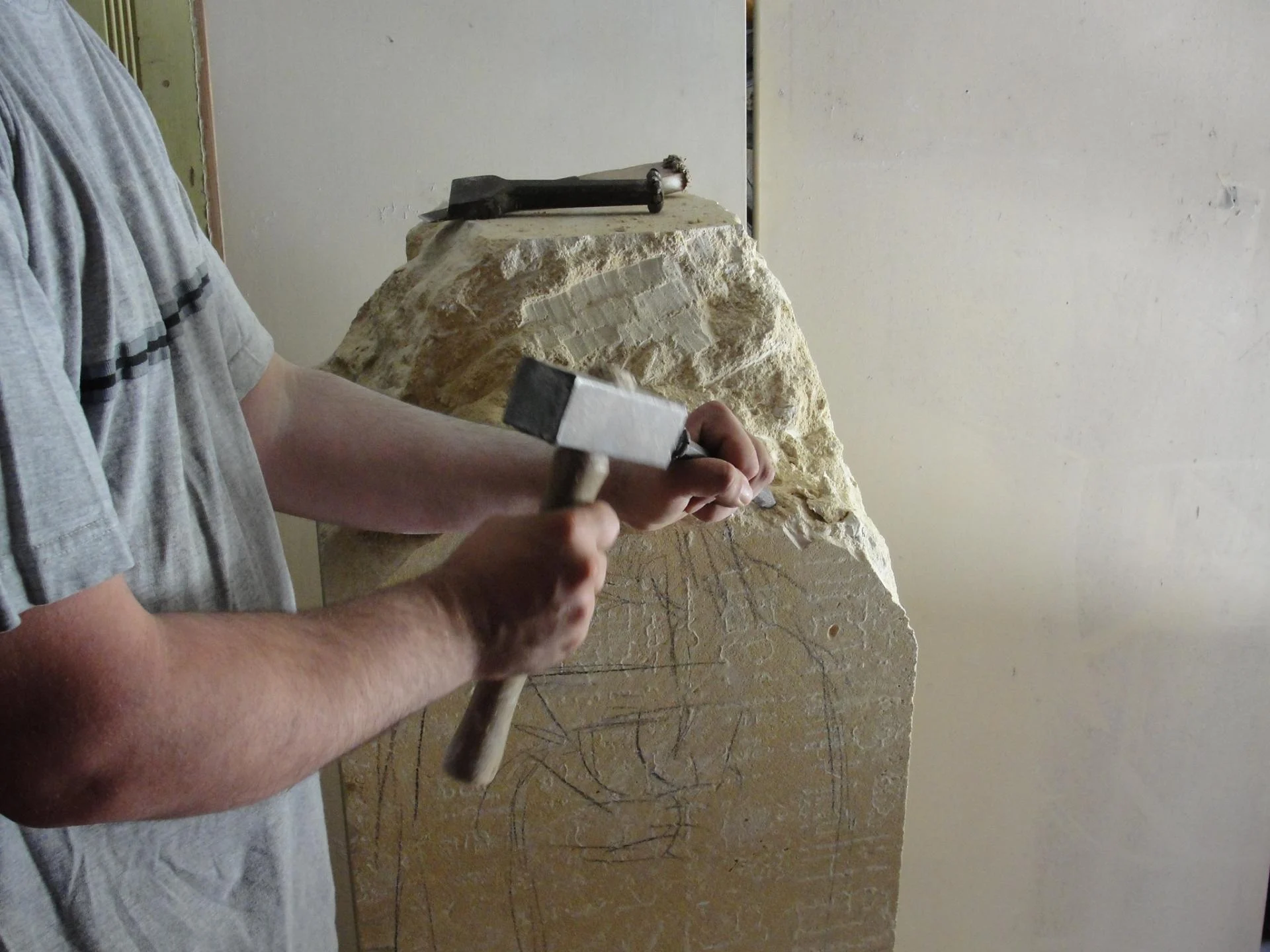 L'artiste au travail sur la statue Saint Jean — le sculpteur Jean-Joseph Chevalier taillant directement le bloc de pierre à la massette et à la chasse, le dessin reporté visible sur la pierre. La taille directe en plein geste, sans moulage. Sculpture d'art sacré — abbaye de Lagrasse 2016.