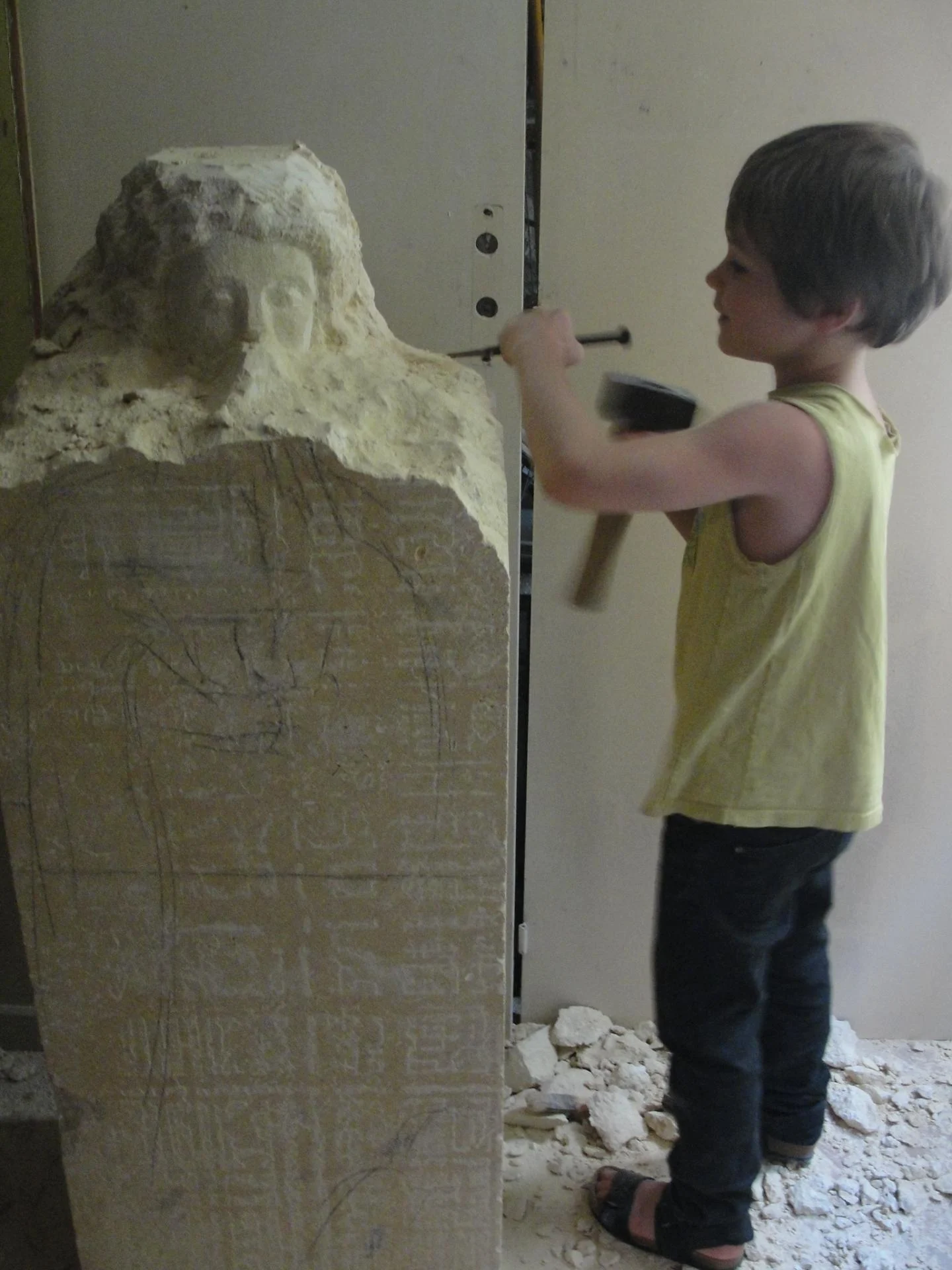 Détail de la statue Saint Jean — le visage en gros plan, tête émergeant de la pierre encore brute, lumière rasante. Le disciple bien-aimé recueilli, qui mène au silence. Pierre naturelle 1,20 m, taille directe, non finito. Statue de saint en pierre véritable, sculpture religieuse d'art sacré — abbaye de Lagrasse, atelier Jean-Joseph Chevalier.