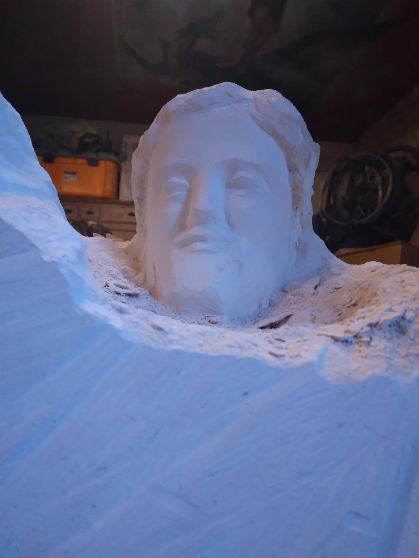Vue d'atelier en gros plan : un visage de la statue Saint Joseph à l'Enfant émerge encore de la matière brute, façonnage au ciseau et à la gradine sur pierre calcaire, atelier de Brignoles, sculpteur d'art sacré Jean-Joseph Chevalier