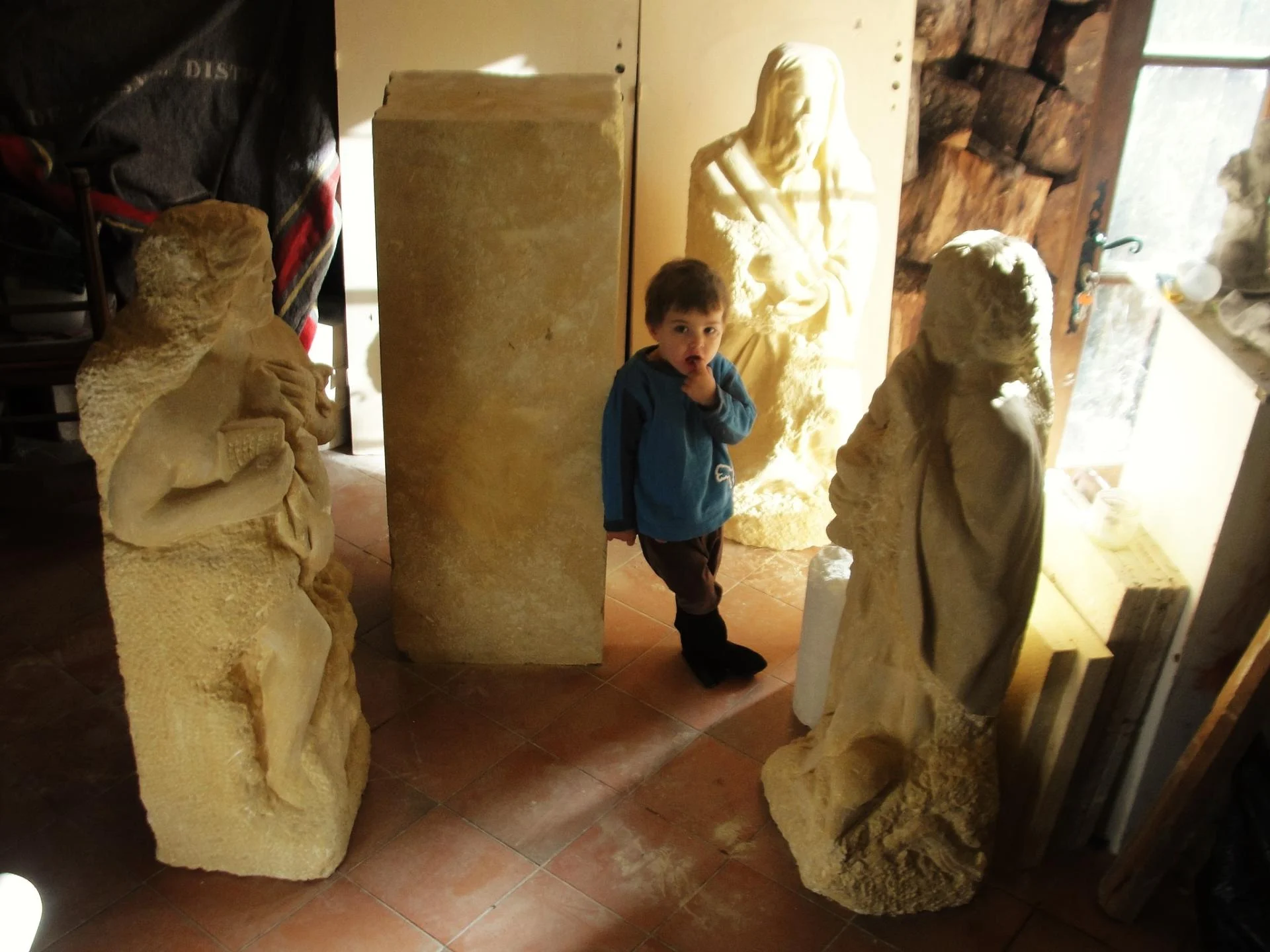 L'atelier — la statue Saint Luc et un autre évangéliste du tétramorphe vus à l'échelle humaine, un jeune enfant près des figures de pierre et des blocs. Pierre calcaire naturelle 1,20 m, taille directe. Ensemble des quatre évangélistes pour l'abbaye de Lagrasse 2016 — Jean-Joseph Chevalier, sculpteur d'art sacré chrétien (Var).