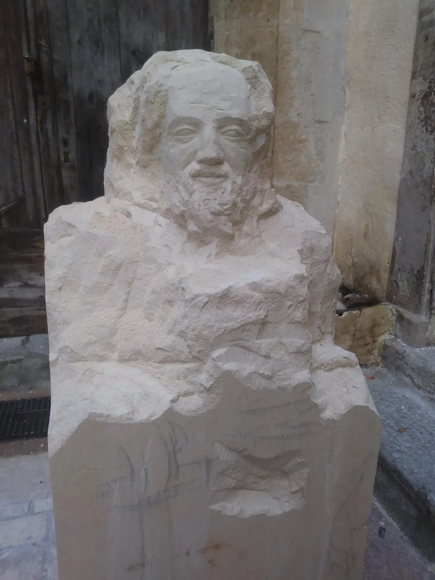 Statue Saint Paul en cours de dégrossissage à la pointe — buste, visage barbu et épaule émergeant du bloc de pierre calcaire encore brut. Taille directe par un sculpteur sur pierre. Statue religieuse 140 cm pour l'abbaye Saint-Paul de Wisques. Sculpteur d'art sacré Jean-Joseph Chevalier.